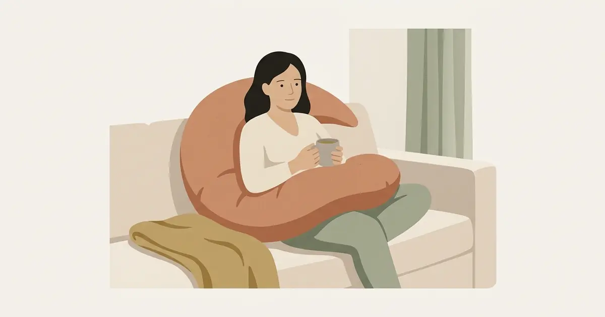 A woman rests on a sofa with a crescent pillow around her waist and a mug of tea in hand