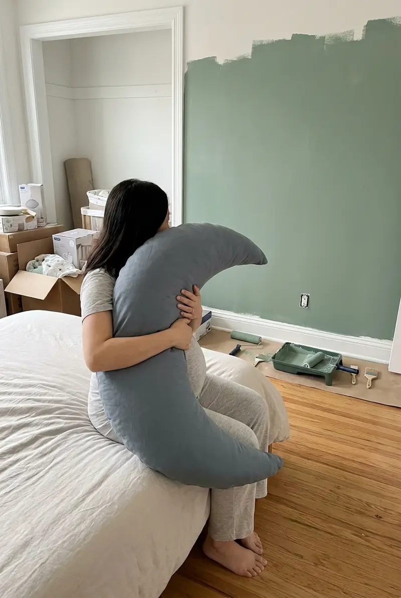 An expecting mother hugging The Cove Pregnancy Pillow in Slate, a half painted nursery behind