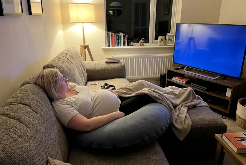 An expecting mother lying on a sofa with The Cove Pregnancy Pillow in Slate